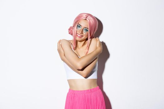 Portrait of beautiful and tender young woman celebrating halloween in glamorous outfit with pink wig, embracing her body and smiling at camera, standing over white background.