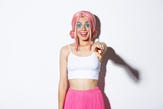 Portrait of excited happy young girl in pink wig and halloween costume, pointing finger at camera, showing something awesome, standing over white background.