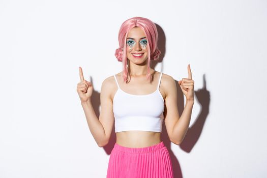 Portrait of happy smiling female model with party wig and glamour halloween makeup, pointing fingers up, showing your logo, standing over white background.