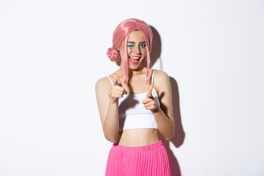 Sassy attractive girl in halloween costume and pink wig, pointing at camera and smiling, congratulating or praising someone, standing over white background.