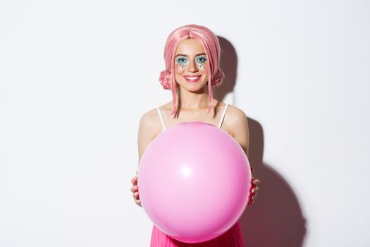 Image of beautiful glamour girl in pink wig, holding big balloon and smiling, celebrating holiday, standing over white background.