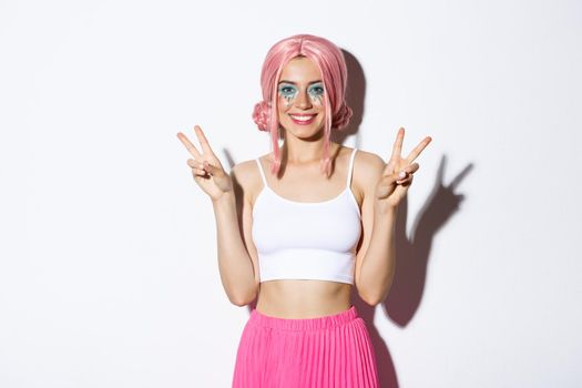 Beautiful smiling woman celebrating halloween in pink wig, showing peace signs, standing over white background.