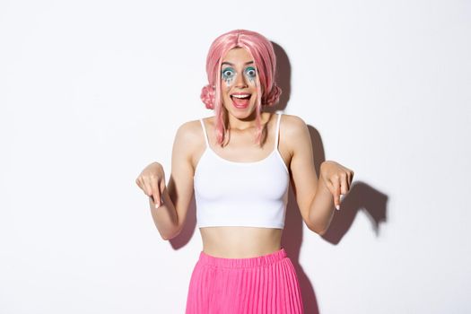 Portrait of excited party girl in pink wig and halloween outfit, looking amused and happy while pointing fingers down, standing over white background.