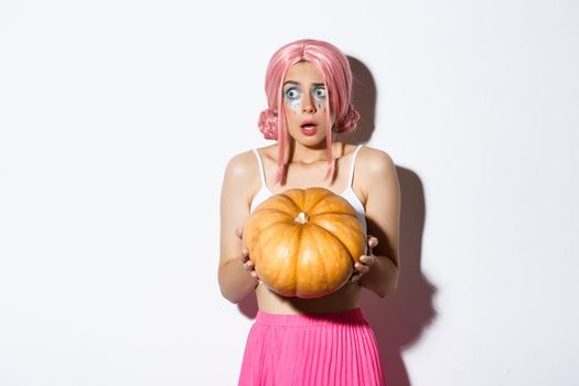 Portrait of confused and scared girl with pink wig and bright makeup, holding pumpkin for halloween and looking left, standing over white background.