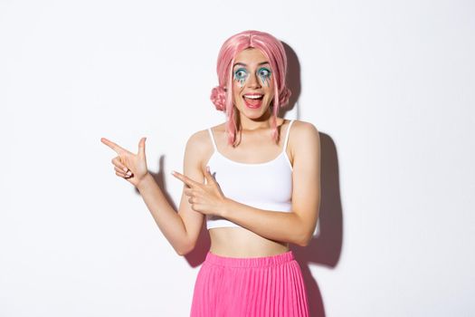 Image of enthusiastic party girl in pink wig and halloween makeup, looking at something fantastic, pointing fingers left, standing over white background.