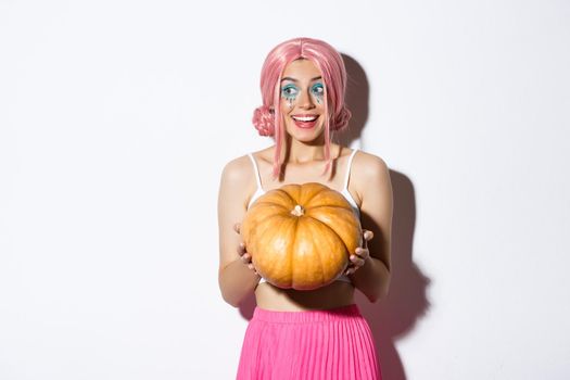 Portrait of attractive girl in pink wig looking amused and smiling, holding pumpkin for halloween party, standing over white background.