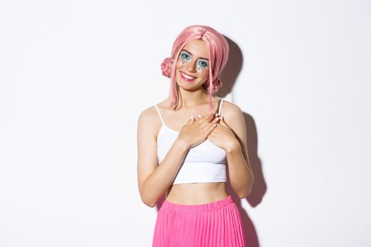 Image of cute party girl in pink wig looking thankful, holding hands on heart and smiling satisfied, standing against white background.
