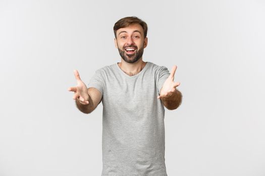 Handsome bearded caucasian man in basic grey t-shirt, stretching hands and reaching for a hug, smiling happy, standing over white background.
