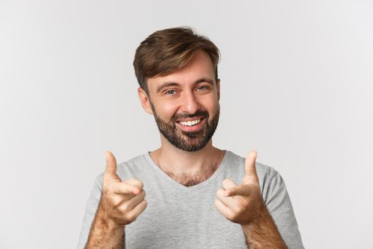 Close-up of handsome adult man with beard, smiling and pointing fingers at camera, standing in gray t-shirt over white background.