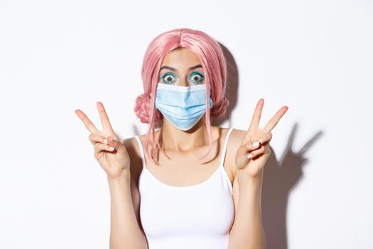 Coronavirus, social distancing and lifestyle concept. Close-up of pretty party girl in medical mask and pink wig, showing peace signs looking excited, white background.