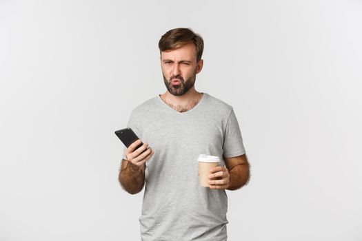 Image of disappointed frowning guy in gray t-shirt, drinking takeaway coffee and holding mobile phone, standing over white background.