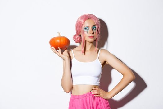 Portrait of sassy attractive girl in pink wig, holding pumpkin for jack-o-lantern, celebrating halloween, standing over white background.