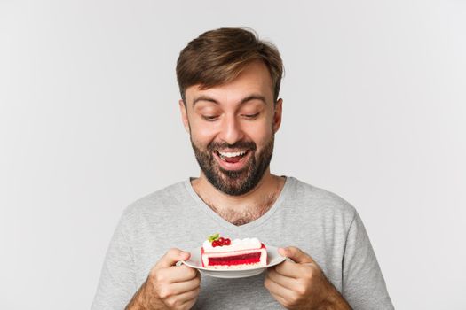 Close-up of handsome smiling man holding cake, standing over white background, excited to eat dessert.
