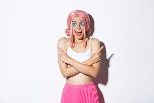 Portrait of excited smiling girl with pink wig and party makeup, pointing fingers sideways but looking left with happy expression, standing over white background.
