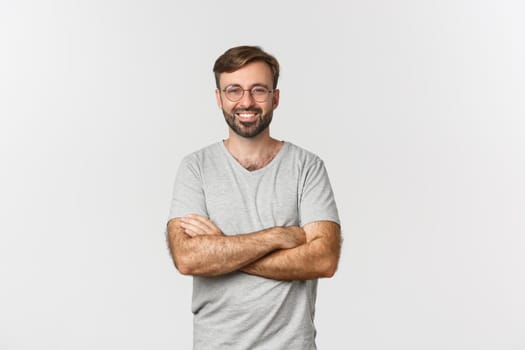 Image of confident handsome man in gray t-shirt and glasses, cross arms on chest and smiling, standing over white background.