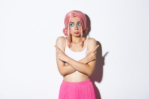 Portrait of clueless silly young girl in pink wig and halloween costume, shrugging and pouting indecisive, pointing fingers sideways, standing over white background.