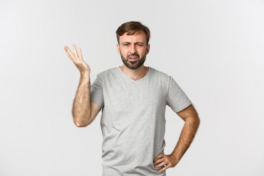 Portrait of confused bearded man in gray t-shirt, hear something stupid and looking skeptical, standing over white background.