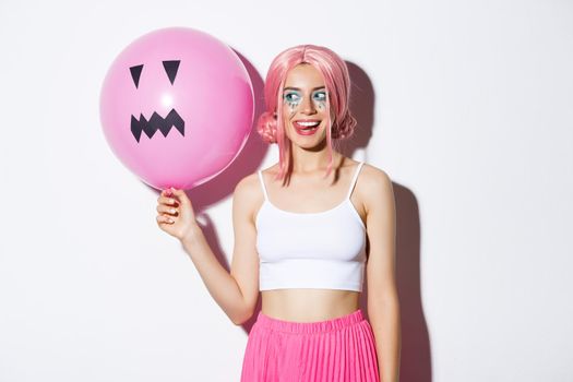 Image of coquettish party girl with bright makeup, wearing pink wig, holding balloon with Jack-o'-lantern face, celebrating halloween.