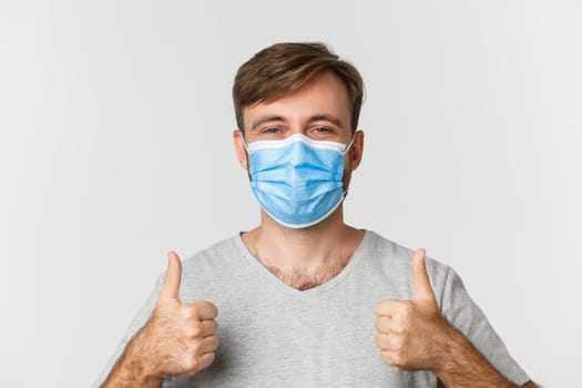 Concept of pandemic, covid-19 and social-distancing. Close-up of cheerful caucasian man in medical mask, showing thumbs-up, standing over white background.