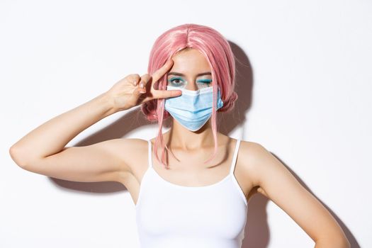 Coronavirus, social distancing and lifestyle concept. Close-up of beautiful cheerful girl in medical mask and pink wig, staying positive, showing peace sign and winking joyfully.