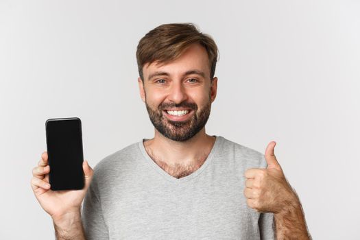 Close-up of smiling handsome man in gray t-shirt, showing mobile phone screen and thumbs-up, recommending app, standing over white background.