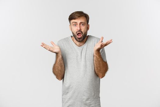 Portrait of confused and worried bearded guy, wearing casual t-shirt, looking at something complicated and shrugging, standing over white background.