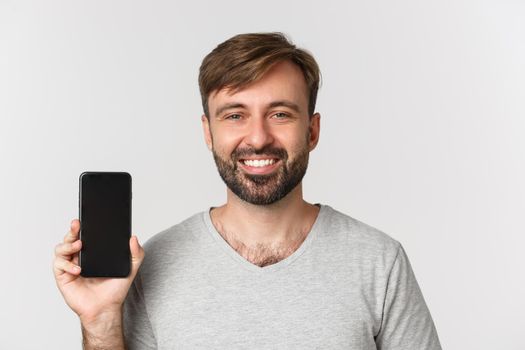 Close-up of smiling handsome man in gray t-shirt, showing mobile phone screen, recommending app or shopping site, standing over white background.