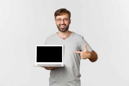 Handsome caucasian guy with beard, wearing gray t-shirt and glasses, pointing at laptop screen, showing something online, standing over white background.