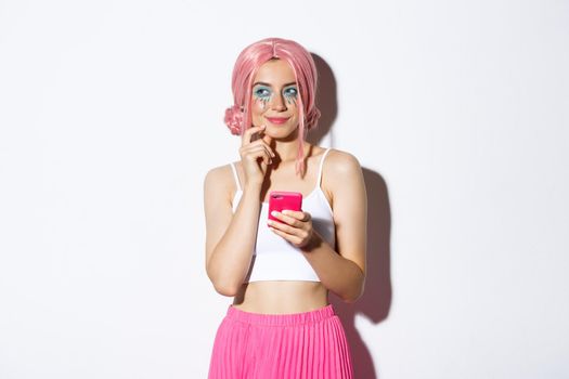 Portrait of thoughtful girl have interesting idea, looking away and thinking while holding smartphone, wearing pink wig for party, celebrating holiday.