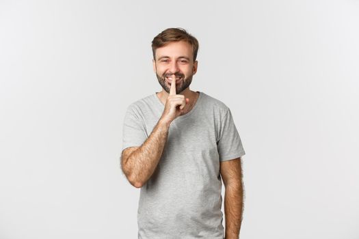 Portrait of handsome smiling man in grey t-shirt, hushing at you kindly asking to keep quiet, standing over white background.