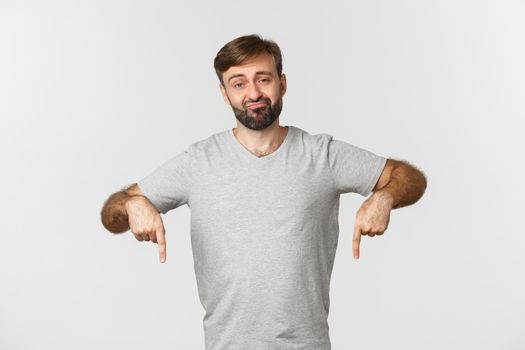 Portrait of unimpressed skeptical guy with beard, wearing gray t-shirt, sulking and pointing fingers down disappointed, standing over white background.