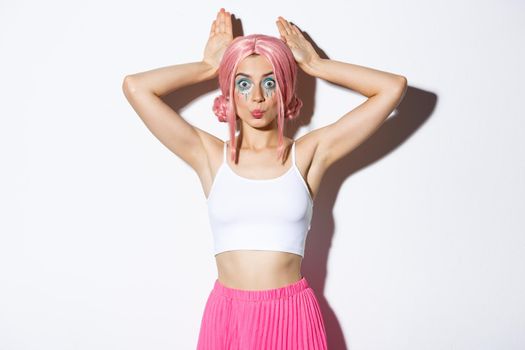 Portrait of lovely party girl in pink anime wig, showing bunny ears and pouting silly, celebrating halloween, standing over white background.