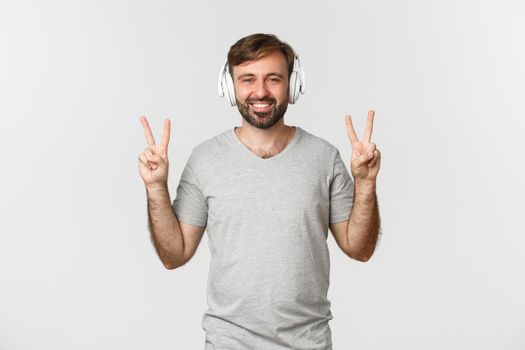 Image of happy caucasian man in gray t-shirt, listening music in headphones and showing peace signs, smiling at camera, standing over white background.