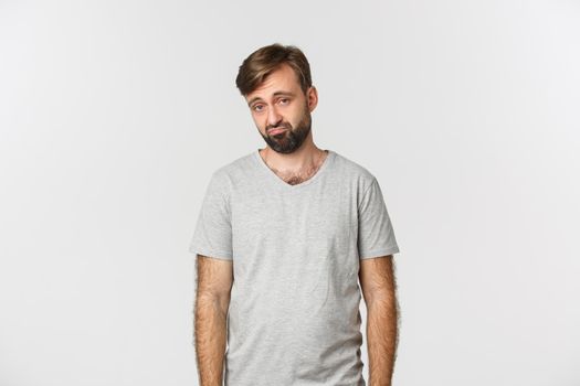 Portrait of sad and gloomy bearded man in gray t-shirt, frowning and looking miserable, standing over white background.