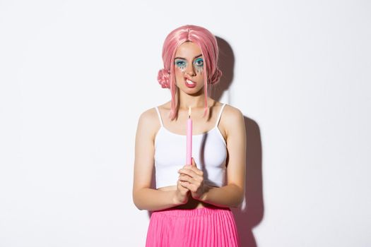 Image of annoyed girl in pink wig, holding candle and showing teeth, celebrating halloween, standing over white background.