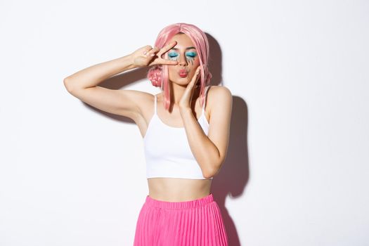 Image of attractive glamour girl in pink wig, bright halloween makeup, standing over white background with peace sign and closed eyes, kissing someone.
