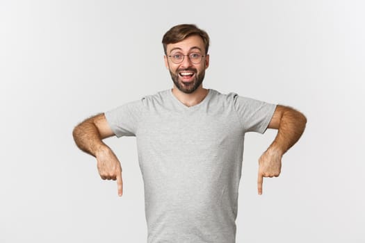 Cheerful bearded man smiling, pointing fingers down, showing logo, wearing gray t-shirt, wearing gray t-shirt, standing over white background.