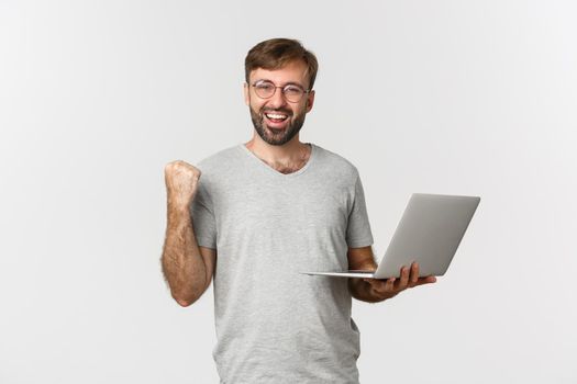 Portrait of cheerful handsome guy in glasses and gray t-shirt, holding laptop, achieve goal, winning something, standing over white background.