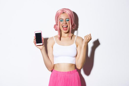 Image of beautiful party girl rejoicing, celebrating holiday, showing smartphone screen and shouting for joy, wearing pink wig and halloween outfit.
