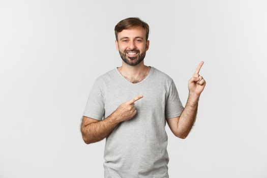 Image of happy handsome guy with beard, wearing gray t-shirt, pointing fingers at upper right corner logo and smiling, standing over white background.