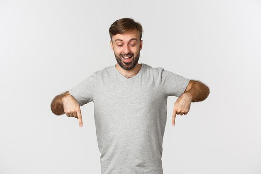 Portrait of excited adult male model in gray t-shirt, looking and pointing down, showing product advertisement, standing over white background.