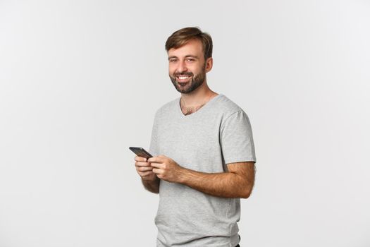 Portrait of handsome bearded guy with a smartphone, smiling pleased at camera, standing over white background.
