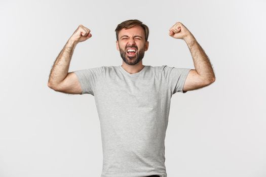 Portrait of happy guy feeling like a champion, raising hands up and rejoicing, shouting for joy, celebrating victory and triumphing, standing over white background.