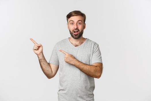 Image of amazed bearded guy in gray t-shirt, pointing fingers at upper left corner logo, showing advertisement, standing over white background.