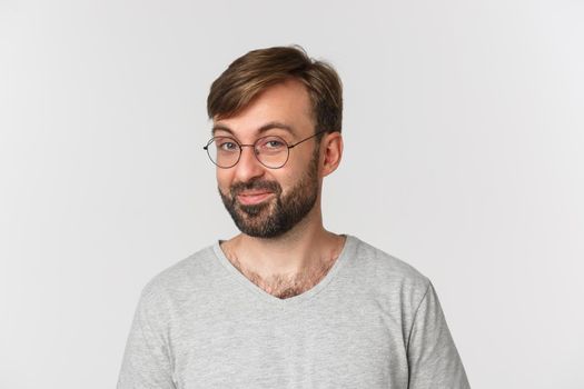 Close-up of curious bearded man in gray t-shirt and glasses, smiling intrigued at camera, standing over white background.