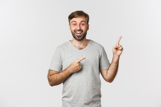 Happy good-looking adult guy with beard, wearing t-shirt, pointing fingers at upper right corner, showing banner, standing over white background.