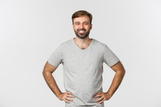Smiling caucasian guy in grey shirt, looking happy and accomplished, holding hands on waist, standing over white background.