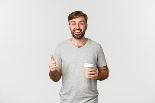 Portrait of cheerful caucasian man in gray t-shirt, drinking coffee and showing thumbs-up, recommend and like cafe, standing over white background.