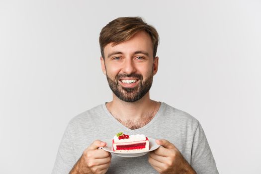 Close-up of handsome smiling man holding cake, standing over white background delighted.