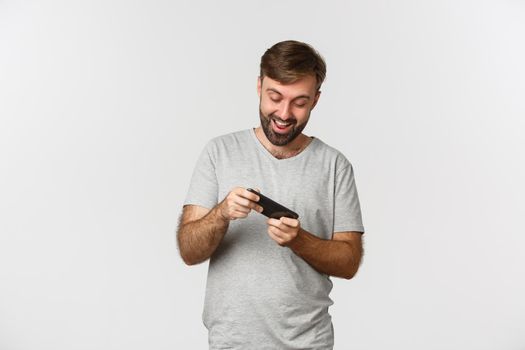 Image of adult bearded man in gray t-shirt, playing mobile games and having fun, standing over white background.